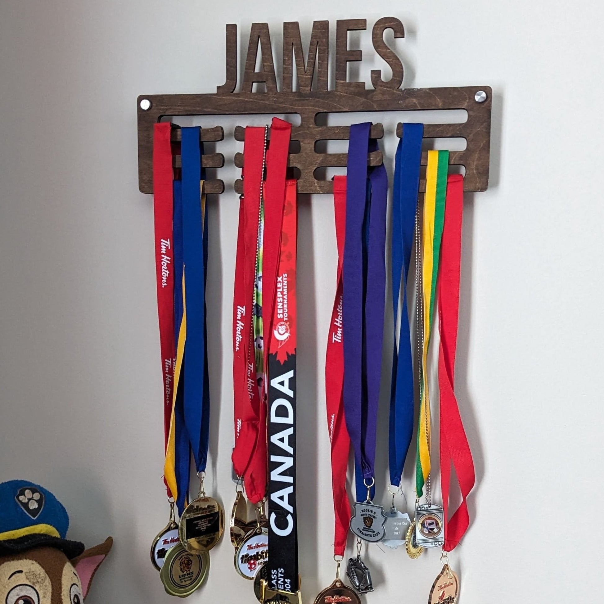 Full picture of a dark walnut stained wooden medal holder customized with the name "James," displaying several hanging medals.