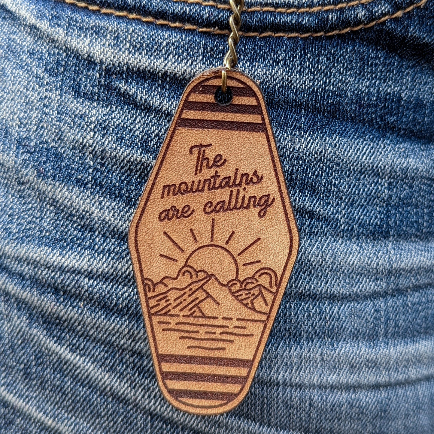 Leather Keychain cut and engraved to look like a vintage motel key with The mountains are calling with an illustration of a sun setting over a mountain engraved on it.